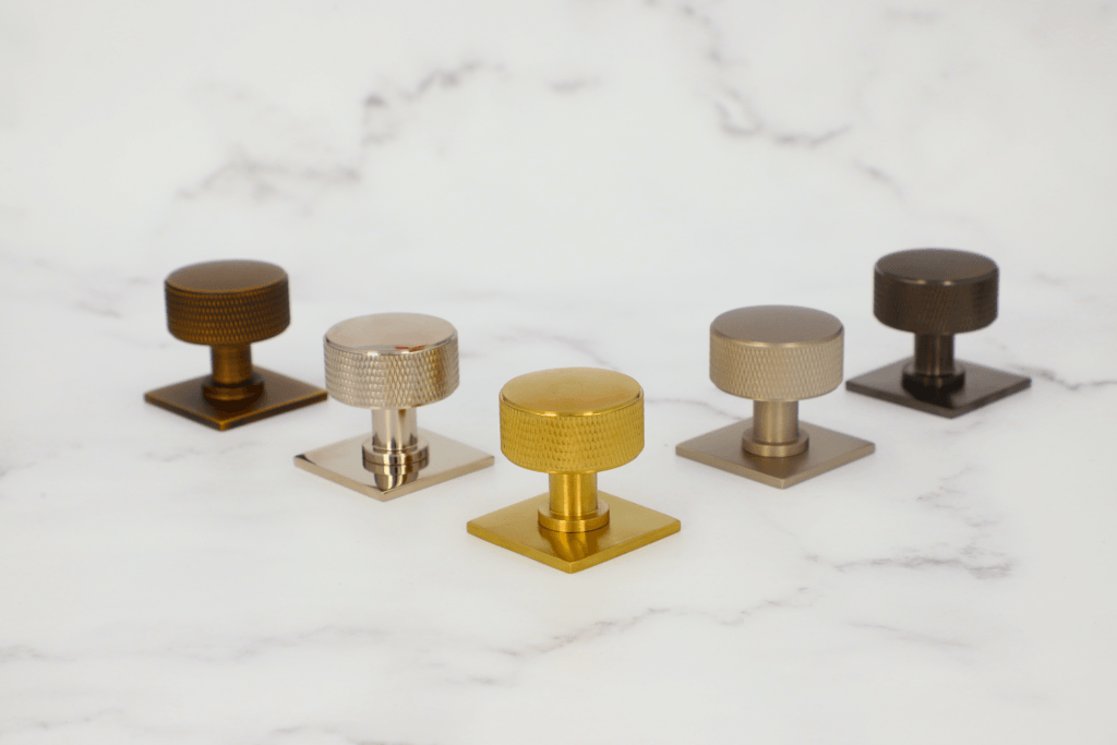 The Alexander & Wilks Lucia knurled cupboard knob shown on the Quantock square backplate in Antique Brass, Polished Nickel, Satin Brass PVD, Satin Nickel and Black PVD against marble backdrop
